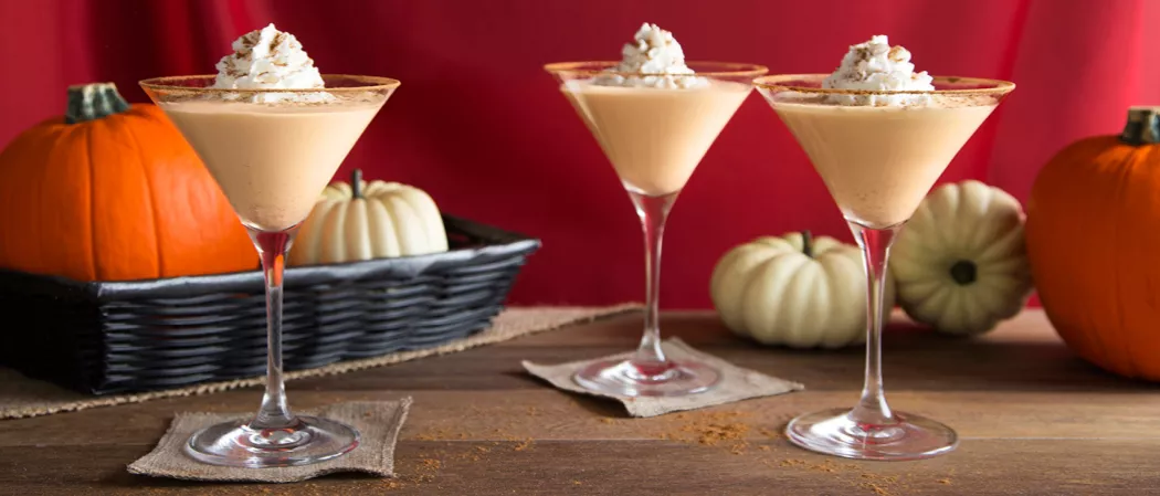Three glasses of Pumpkin Pie Martini, each garnished with whipped cream and Pumpkin Spice, placed on a small piece of cloth. Nearby, a tray of pumpkin on a cloth, with few pumpkins outside the tray.