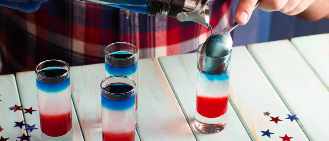 A person pouring blue liquid into glass of Red, White, and Blue Shot using spoon alongside three glass of same drink