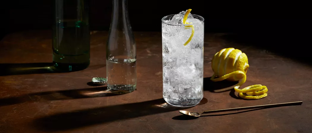 A glass of Roku® Gin Sonic with ice, half-filled water bottle, half-filled green glass, artisticallly chopped lemon, and copper spoon on the table. 