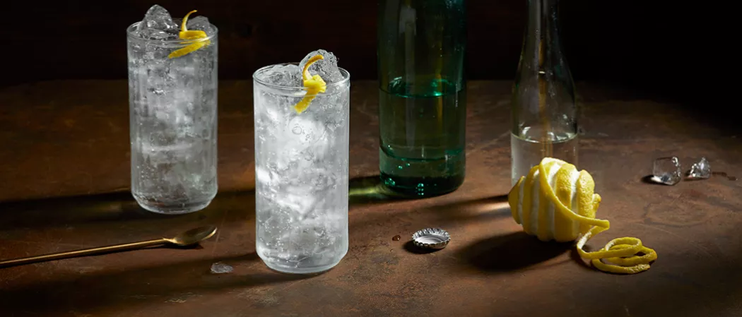 Two glasses of Roku® Gin Sonic with ice, half-filled water bottle, half-filled green glass, artisticallly chopped lemon, and copper spoon on the table. 