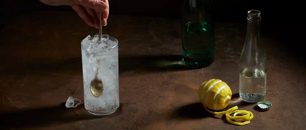 A person muddling copper spoon inside the glass of Roku® Gin Sonic with ice, half-filled water bottle, half-filled green glass, artisticallly chopped lemon, and copper spoon on the table
