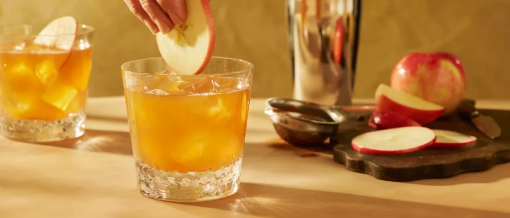 A person garnishing a apple slice on a glass of SQRRL® An Apple A Day along with one more glass of SQRRL® An Apple A Day and chopped unchopped apples