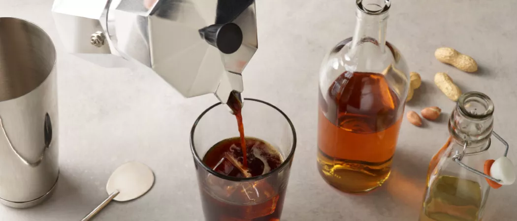A person pouring Espresso into a glass of SQRRL® Carajillo along with SQRRL® Peanut Butter Whiskey bottle and some ground nuts on the kitchen slab
