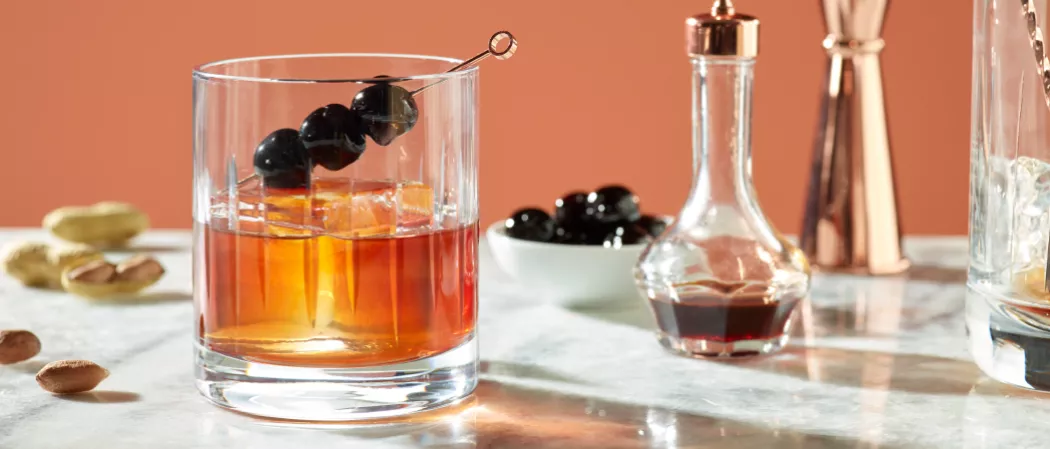 A glass of SQRRL® PB Manhattan  garnished with Maraschino Cherries along with some ground nuts  on the kitchen slab