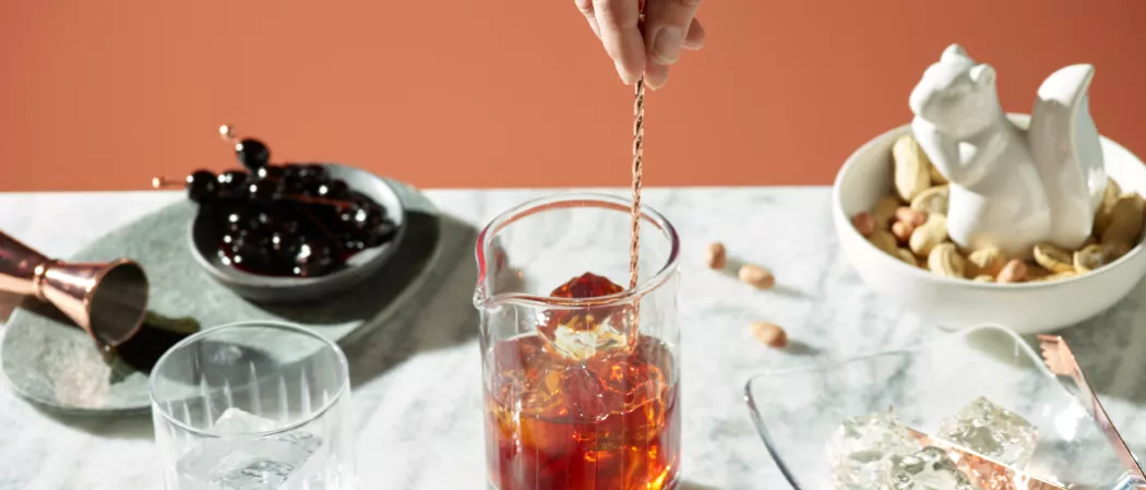 A person stirring ice cubes in a glass of SQRRL® PB Manhattan along with the bowl of Maraschino cherries and a bowl ground nuts with a squirrel statue