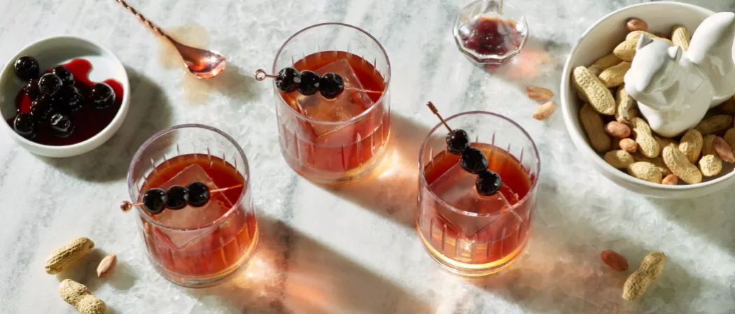 Three glasses of SQRRL® PB Manhattan  garnished with Maraschino Cherries along with some ground nuts in a bowl on the kitchen slab