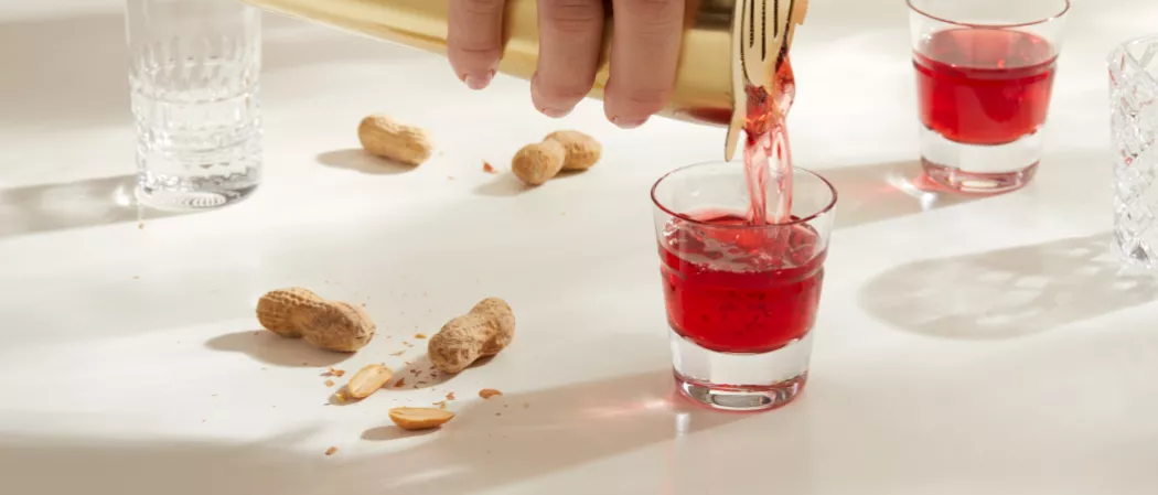 A person pouring DeKuyper® Razzmatazz® Schnapps Liqueur into a glass of  SQRRL® PB&J Shot along with some ground nuts on the kitchen slab