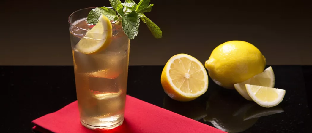 The glass of Sauza® 901® Club, garnished with a lemon slice, mint leaves, and an ice cube, is placed on a red paper alongside  lemon slices and lemon on a black table