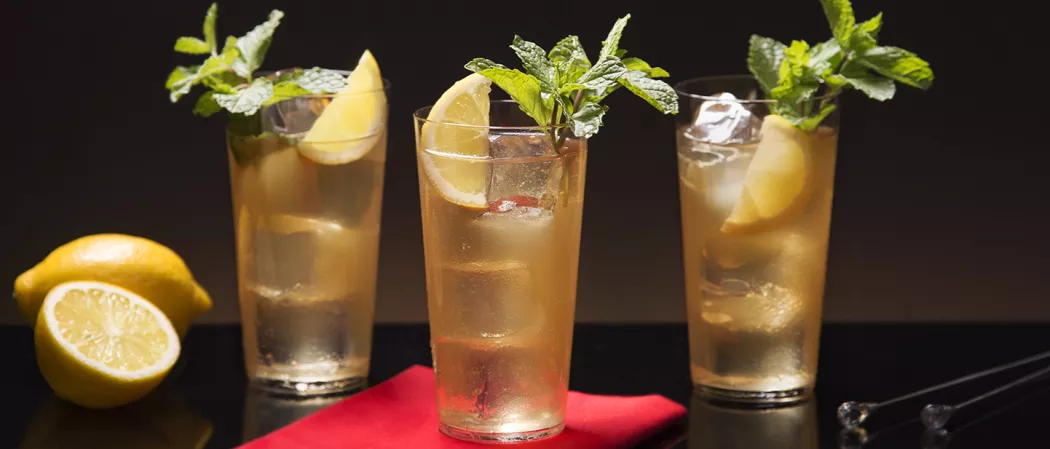 The glass of Sauza® 901® Club, garnished with a lemon slice, mint leaves, and an ice cube, is placed on a red paper alongside two other glasses of same accompanied by lemon slices on one side and a spatula on the other