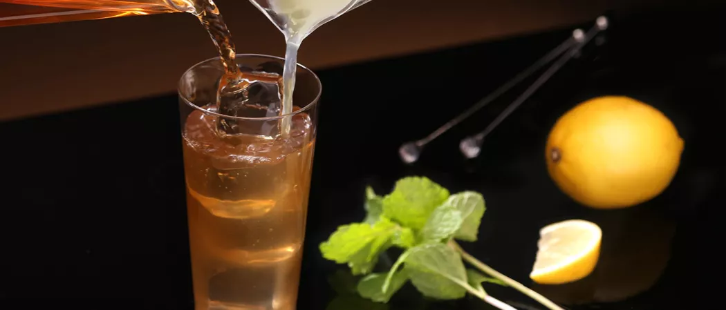 A person is stirring red and white liquid over ice in a glass of Sauza® 901® , alongside mint leaves, lemon, a slice of lemon, and a spatula on a black table