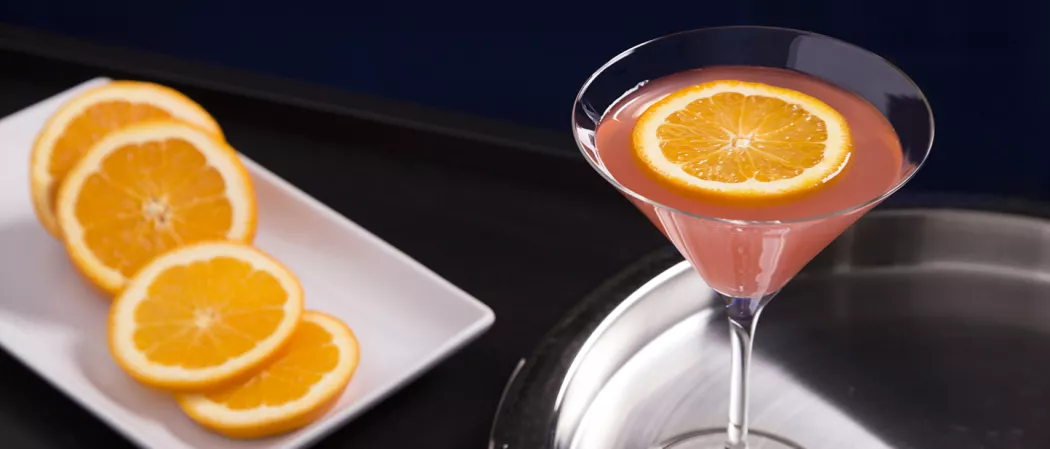 Glass of El Bombín garnished with an orange slice on a steel tray, alongside some additional orange slices on a white tray