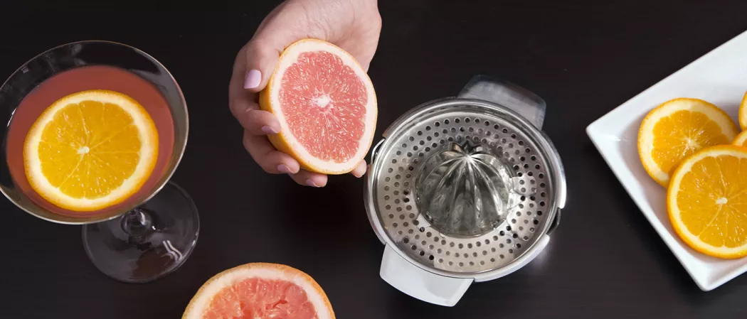 a person holding grapefruit alongside  a glass of EI bombin garnished with orange slice, a juicer and few orange slices on a white tray