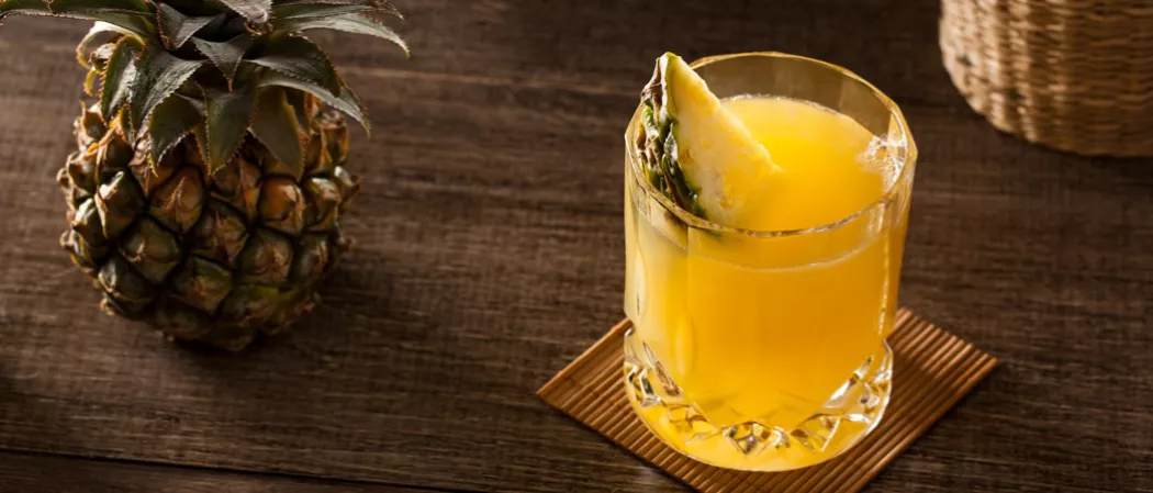 A glass of Pequeña Colada, garnished with a slice of pineapple, sits alongside a pineapple on a wooden table