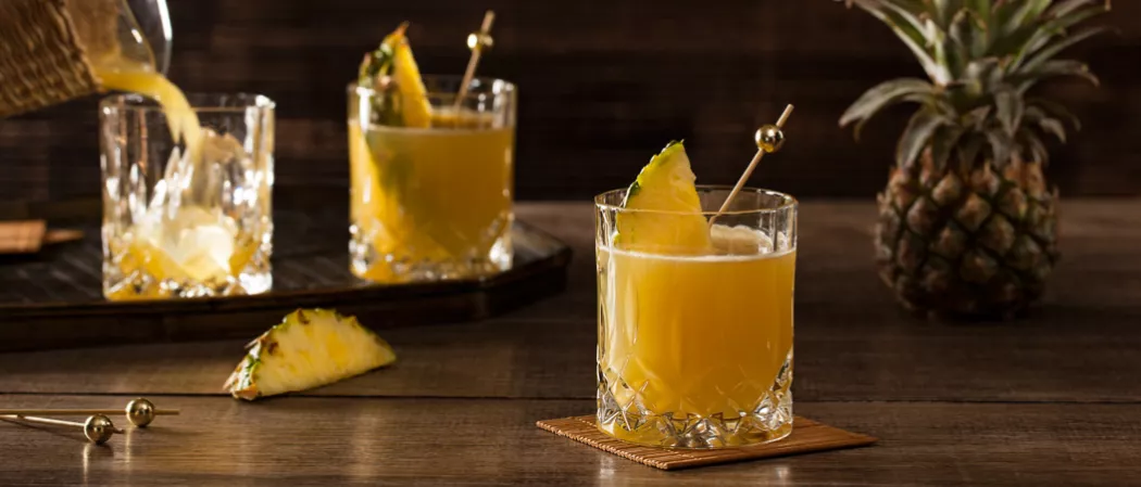 Three glasses of Pequeña Colada, garnished with a slice of pineapple and a wooden toothpicks cocktail stick, one on a cardboard, one is being poured with Pequeña Colada