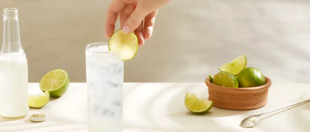 A person garnishing lemon on a glass of Sauza® Mexican Paloma along with a bottle of Grapefruit Soda and a bowl of lemons