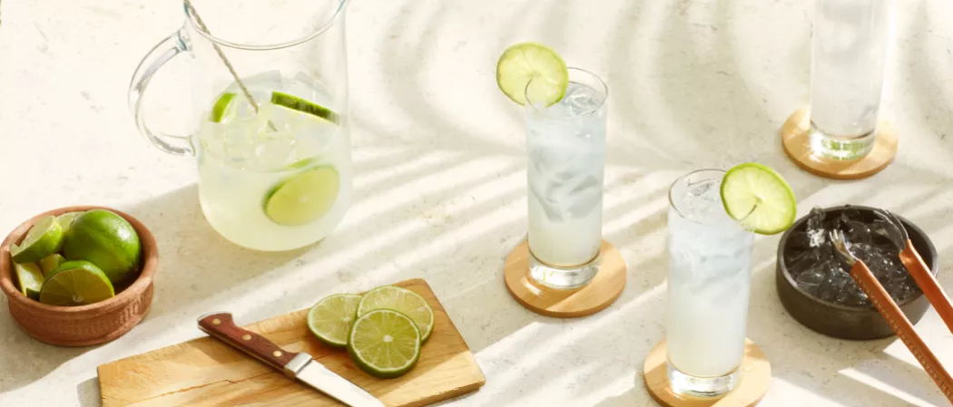 Two glasses and one jar of Sauza® Mexican Paloma garnished with Lime Wheel along with chopped lemons, knife and a small bowl of lemons