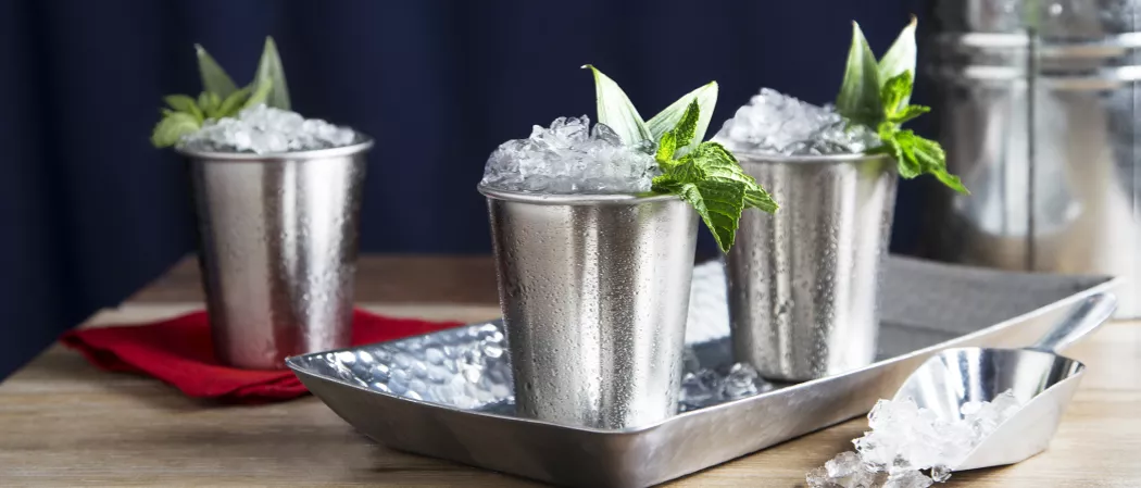 Three glasses of Mexican Julep, each garnished with crushed ice and fresh mint sprigs, one glass is resting on a red cloth, and the other two are placed in a steel tray. Additionally, crushed ice accompanies them