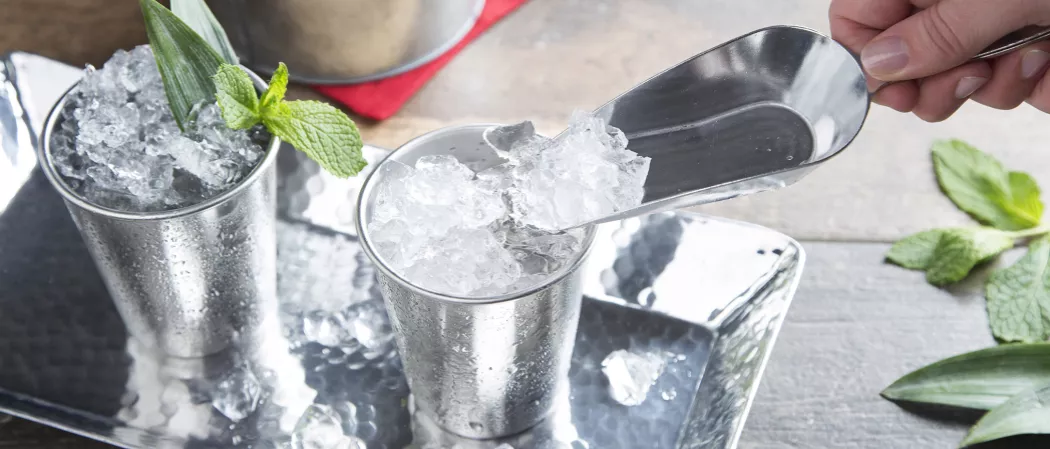 Two pewter mugs of Mexican Julep are placed in a steel tray. One mug is garnished with fresh mint sprigs, while in the other, a person is pouring crushed ice with a spoon