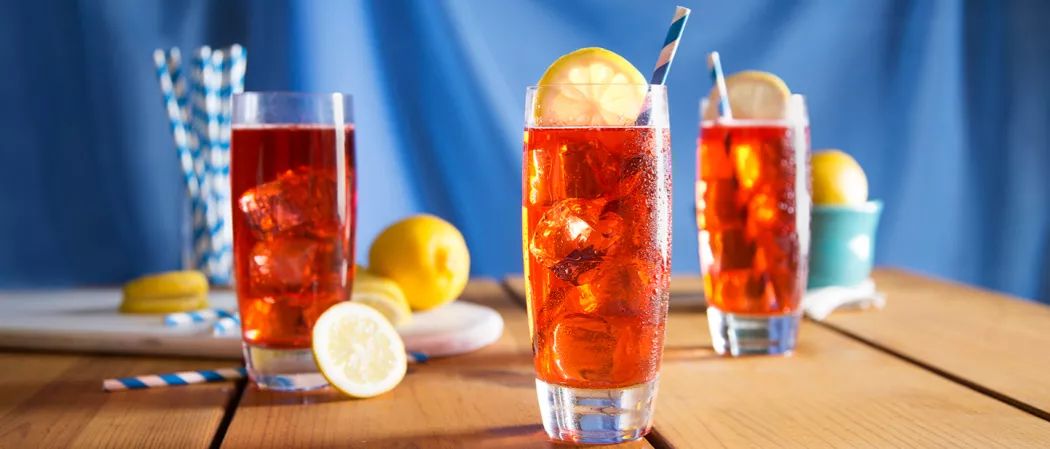 Three glasses of Sloe Gin Fizz, One glass is garnished only with ice cubes, alongside a lemon ,lemon slicse and a straws, other two glasses are garnished with ice cubes, lemon slices, and straws