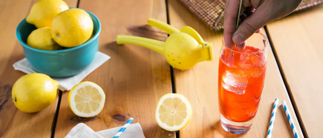 A person is mixing Sloe Gin Fizz, garnished with ice cubes, using a stirring stick, alongside a lemon squeezer, a lemon, and a few lemon slices, all placed on a wooden table. and  a bowl of lemons on a white cloth