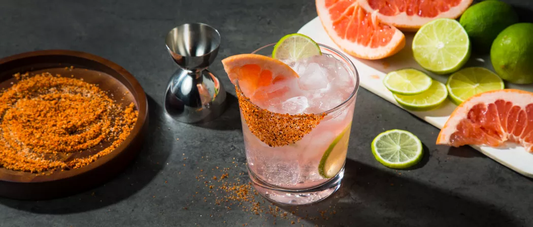 A glass of El Tesoro® Paloma garnished with Grapefruit Wedge ,Salt and Spice Rim and a chopped lemon along with  plate of  salt & spice and chopped grapefruits & lemons