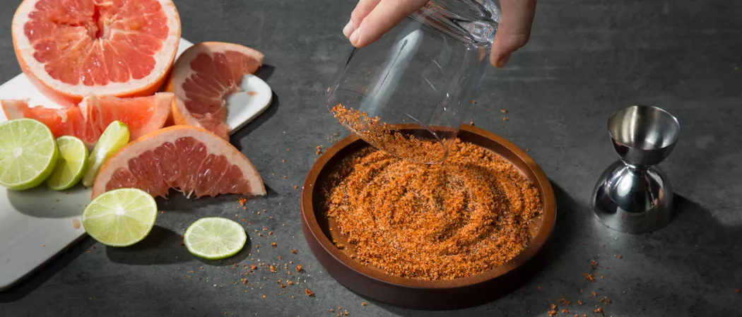 A person holding a glass and dipping it into Salt and Chili Powde on a plate anlong with chopped grapefruite and  lemons