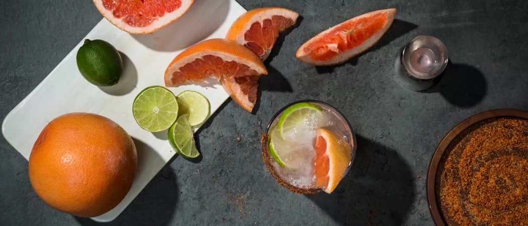 A glass of El Tesoro® Paloma garnished with Grapefruit Wedge ,Salt and Spice Rim and a chopped lemon along with  plate of  salt & spice and chopped grapefruits & lemons