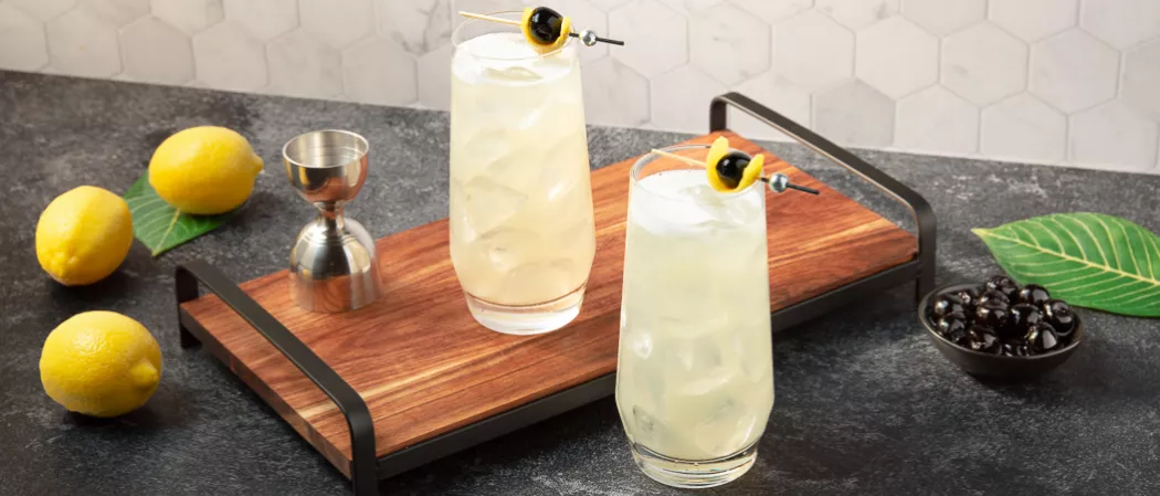 Two glasses of Tokyo Collins garnished with lemon peel and black cherries in a wooden tray on the kitchen slab along with  a jigger, a small bowl of black berries , three lemons 