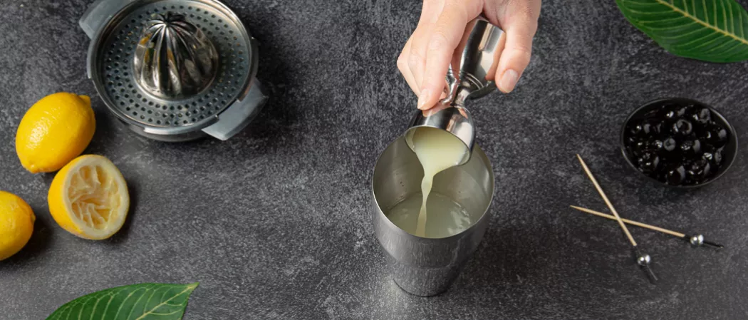A person pouring a lemon juice into glass from a jigger along with a small bowl of black cherries , lemons and a manual juicer cap.