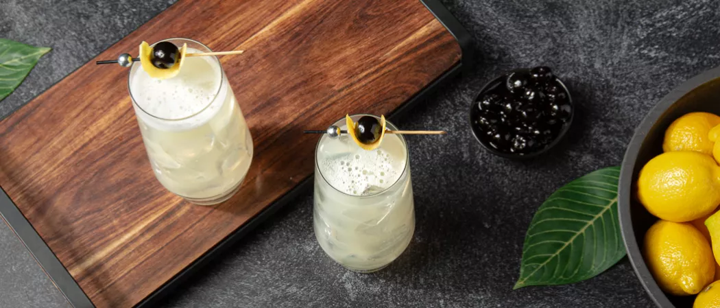Two glasses of Tokyo Collins garnished with lemon peel and black cherries on a wooden try on the kitchen slab along with  two bowls of black berries and lemons 
