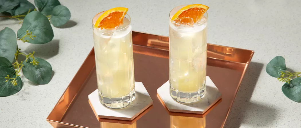 Two glasses of Tom Collins garnished with chopped orange in a copper plate on a stone coaster along with some vibrant leaves 