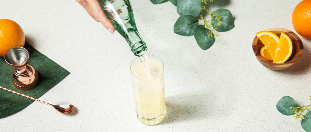A person pouring water in a glass of Tom Collins, alongside a jigger, a copper spoon , oranges placed on green cloth and a bowl of chopped oranges
