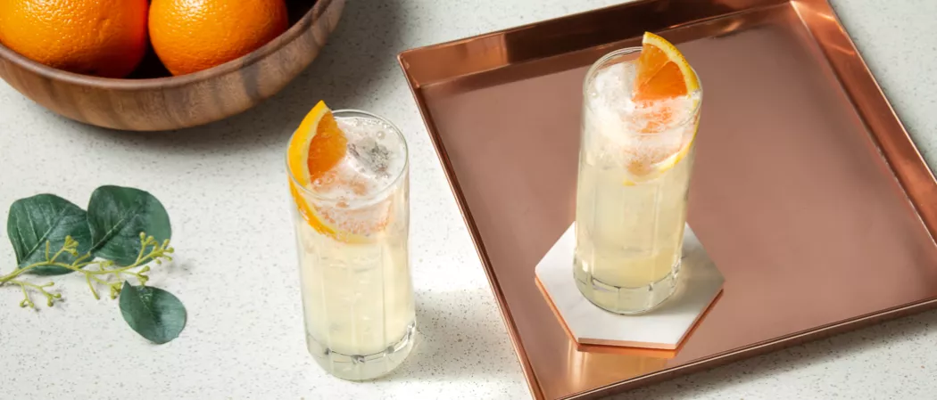 Two glasses of Tom Collins garnished with chopped orange one in a copper plate on a stone coaster and one on the table along with some vibrant leaves 