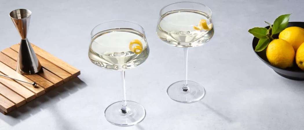 Two glasses of Vesper garnished with lemon peel along with the jigger and a steel spoon on a wooden frame and a bowl of lemons