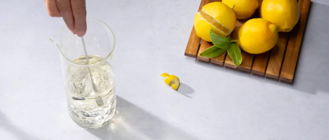 A person stirring a half filled glass jug with Vesper along with few lemons on the wooden board