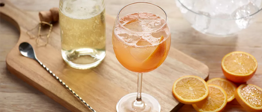 A glass of Maker's Mark® Bourbon Spritz with orange slices on a wooden table along with the silver spoon and a water bottel.
