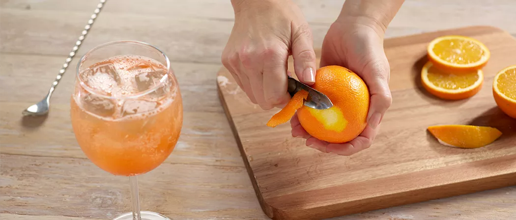  A glass of Maker's Mark® Bourbon Spritz and a person peeling orange along with the silver spoon and chopped orange pieces.