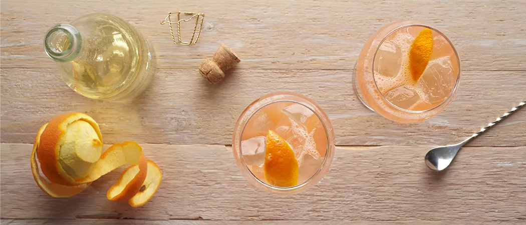 Two glasses of Maker's Mark® Bourbon Spritz garnished with orange peel along with peeled orange ,water bottle and silver spoon