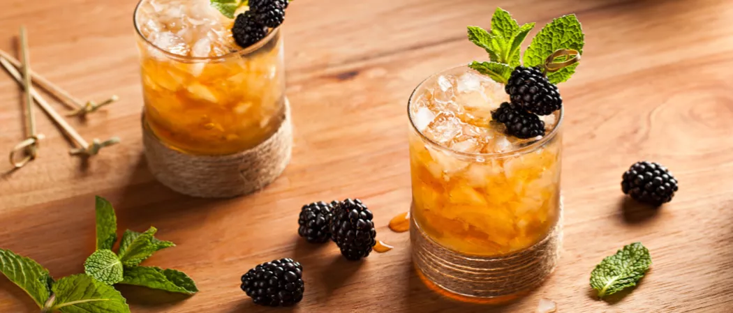 Two glasses of Hats Off Julep on a wooden table garnished with blackberries and mint leaves