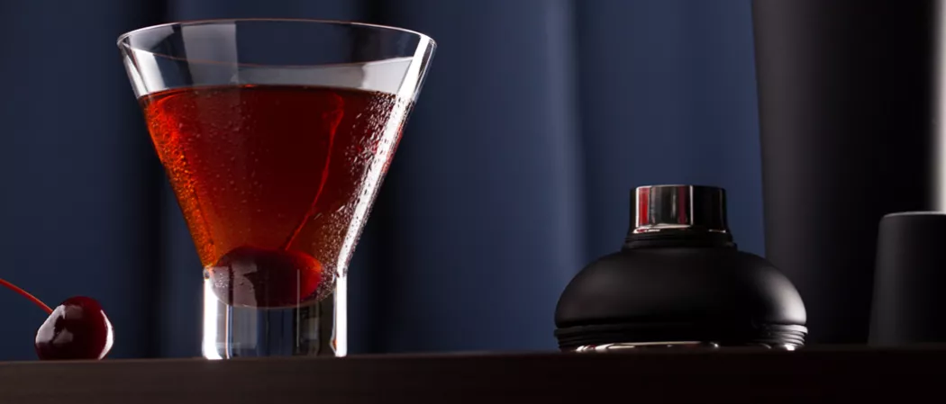 Glass of Jim Beam Black Manhattan garnished with a cherry inside, alongside a cherry and the cap of the shaker