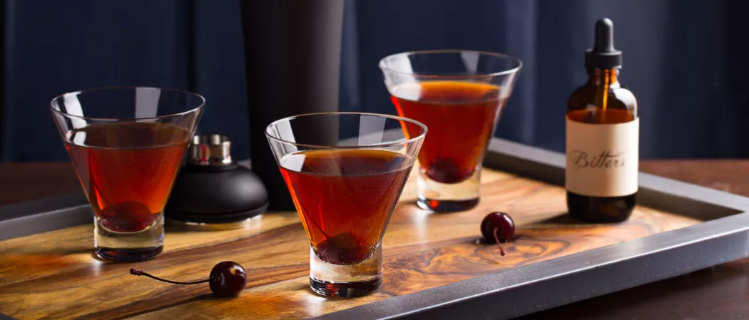 Three glasses of Jim Beam Black Manhattan garnished with a cherry inside, alongside cherries, the cap of the shaker, and a bottle of bitters on a wooden tray