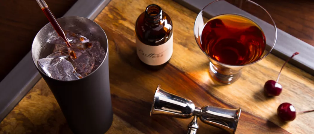 Glass of Jim Beam Black Manhattan next to a bucket of crushed ice, cherries, a bottle of bitters, and a jigger on a wooden tray