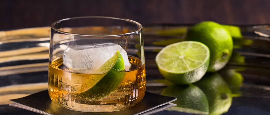 a glass of C.C.® & Ginger garnished with ice and lime slice on coaster alongside few limes