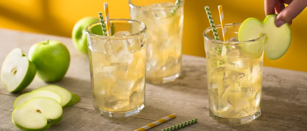 Person garnishing glass of Crisp Apple Ade with apple slice alongside two other glasses of Crisp Apple Ade