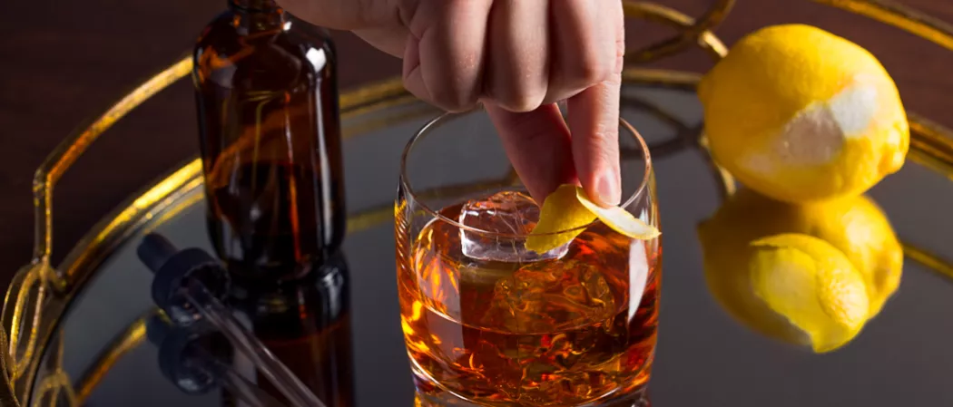 Person garnishing glass of Dry Rye Manhattan garnished with lemon peel alongside  lemon