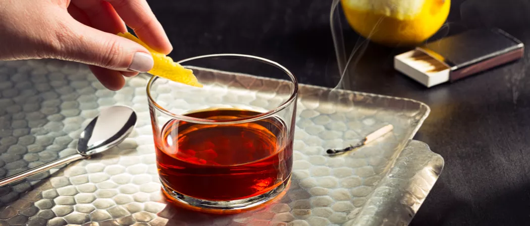 A person garnishes a Hornitos® Black Barrel® Tequila Manhattan with orange peel on a silver tray next to a spoon and a burnt matchstick.