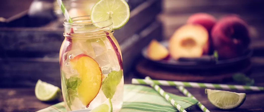 A jar of Peach Daiquiri garnished with lime slice, peach slices, mint leaves and a straws alongside few strawss and lime slices