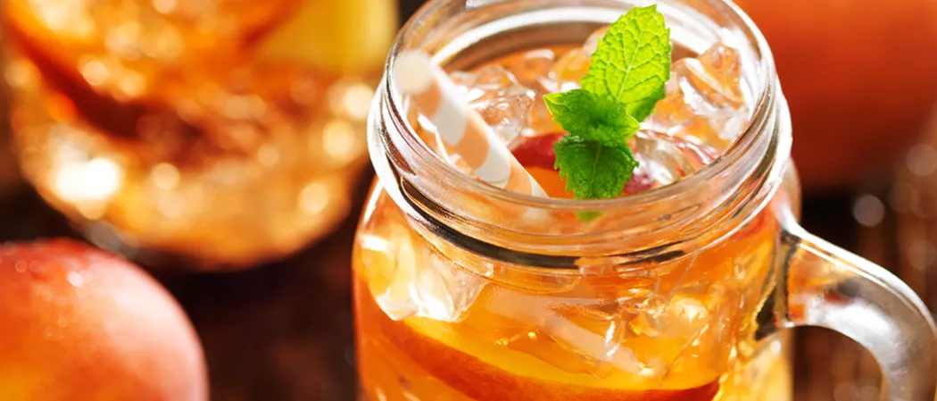 A Glass of Peach Mojito garnished with a mint sprig and a peach slice, with some ice cubes and straws in it.