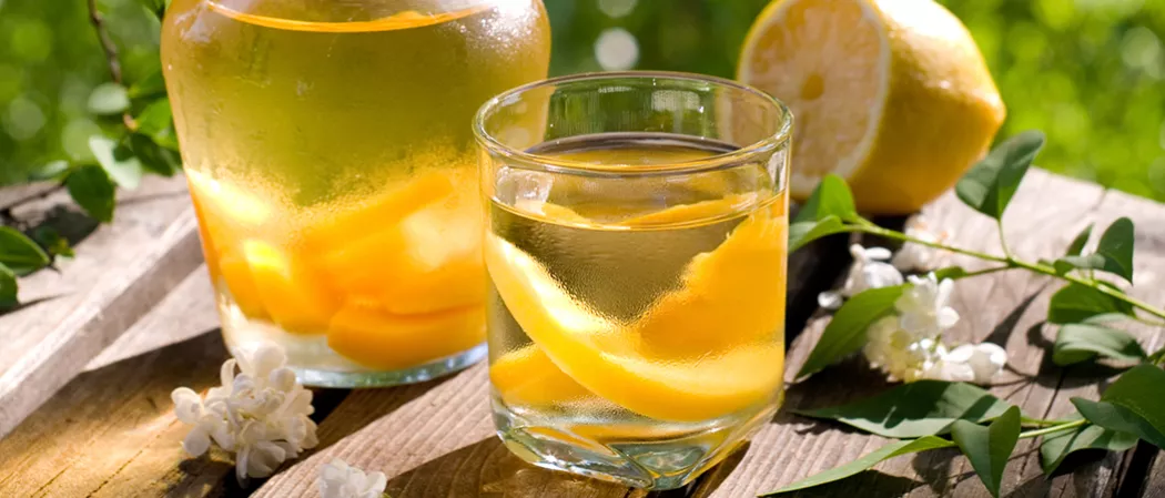 A glass of Cruzan® Peach Sangria with lime slices, a jar filled with the same drink, a halved lemon, and jasmine flowers on the wooden bench.