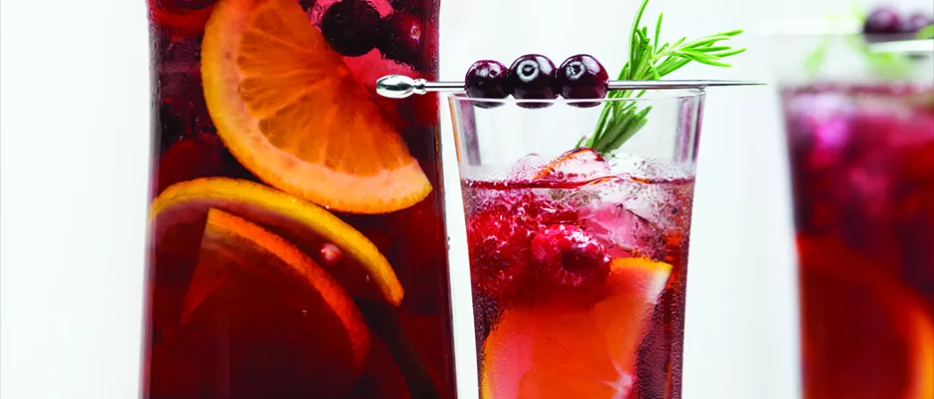 A glass of Razzmatazz® Sangria with ice cubes, garnished with mixed berries, and three mixed berries pierced on a cocktail pick, placed on the rim of the glass.